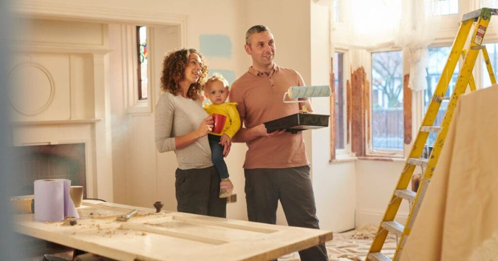 Homeowner holding baby and looking at Painted wall with paint tools in pircture | Building Material Reviews 